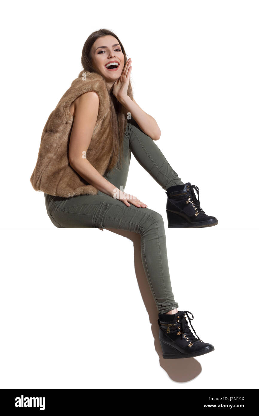 Beautiful young woman in brown fur waistcoat, khaki pants and black boots sitting on a top, holding hand on chin, looking at camera and laughing. Full Stock Photo