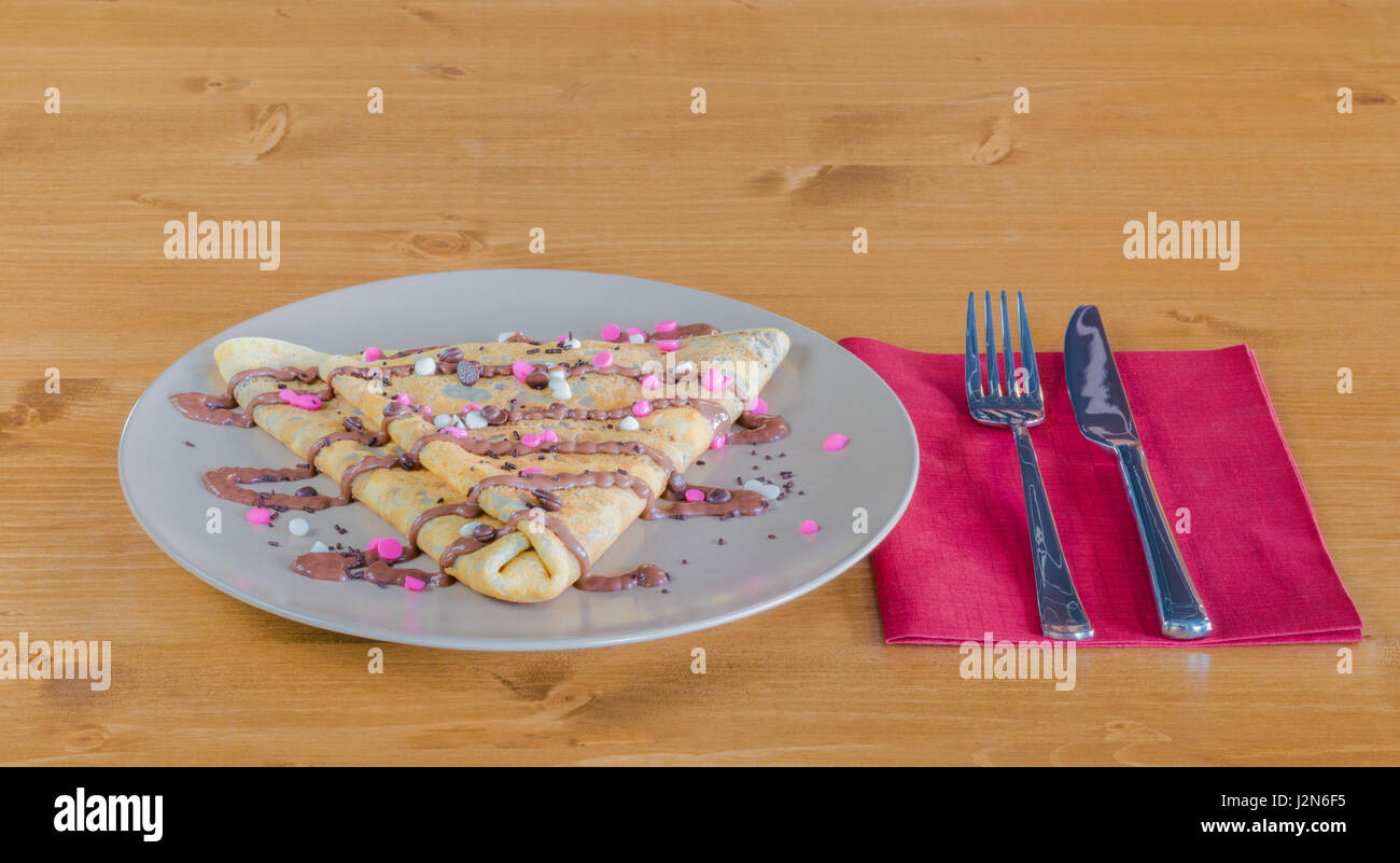 Crepe with chocolate cream and chocolate chips and chocolate topping served on white plate on a wooden table with fork and knife on a red napkin Stock Photo