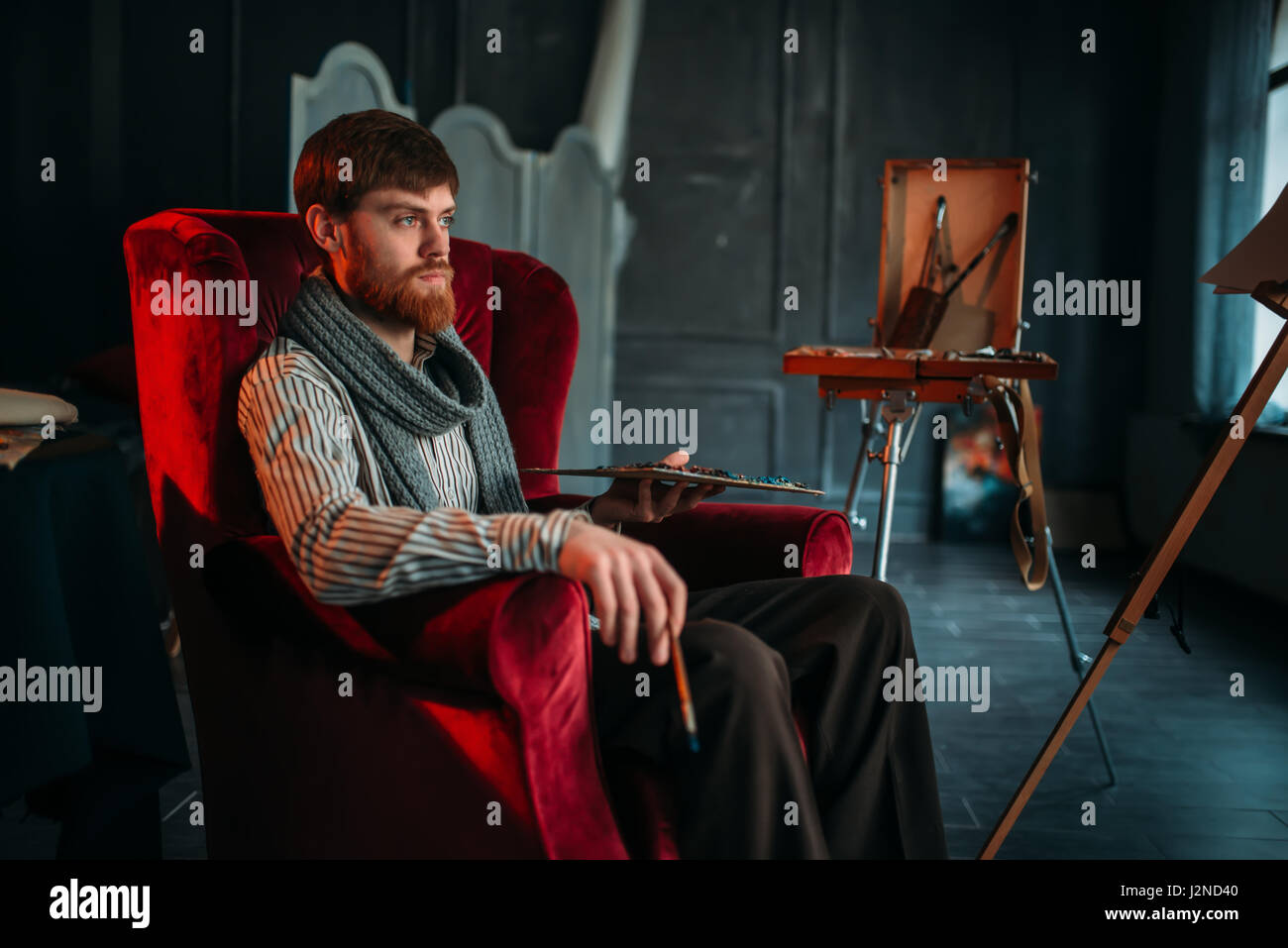 Painter with palette and brush in hand sitting on a chair, art studio on background Stock Photo