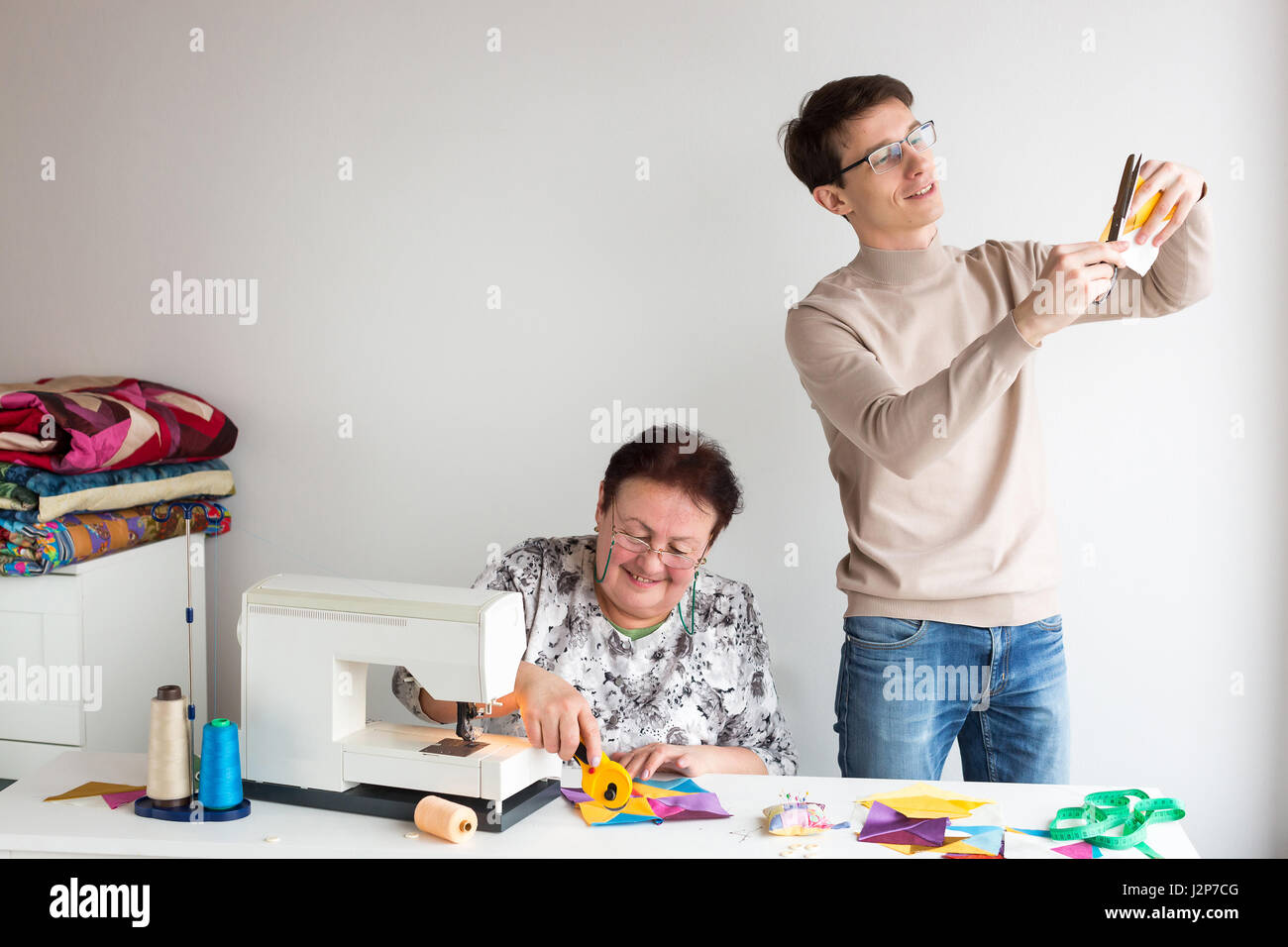 patchwork - the work of the two tailors in the studio - woman cut with scissors colored fabric behind table with fabrics and tools, a handsome man stands by and cuts with scissors patchwork fabric. Stock Photo