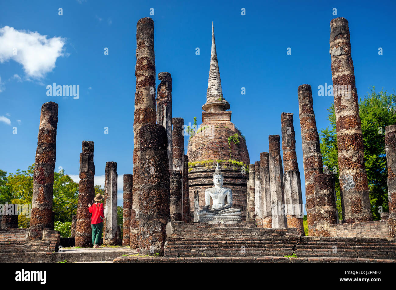 Tourist Woman in red costume with camera looking at ancient Buddha surround by ruined column in Wat Sa Si of Sukhothai Historical Park, Thailand Stock Photo