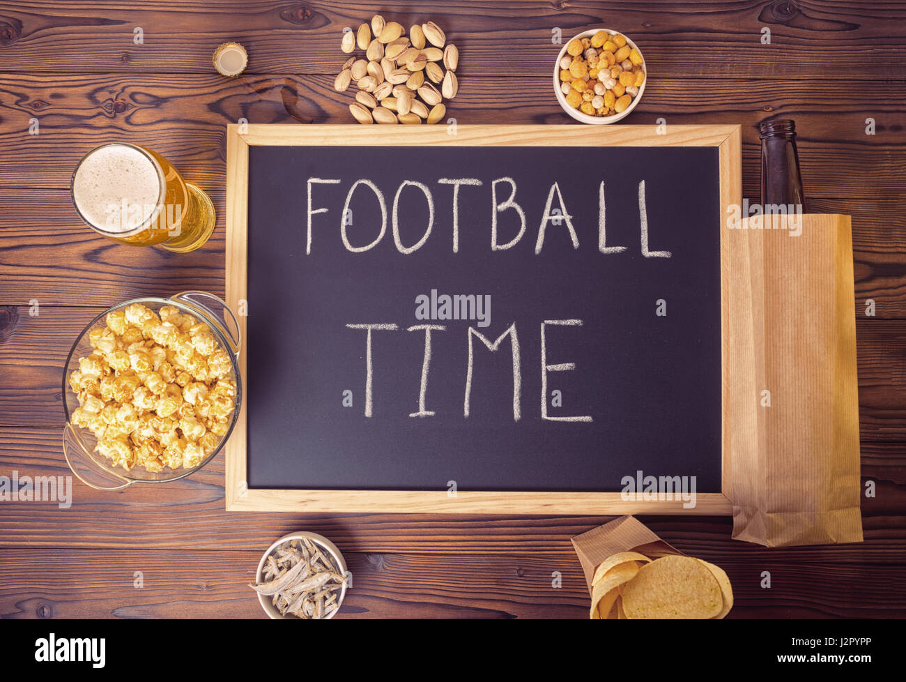 football fans setting of beer bottle in brown paper bag,  glass, chips, pistachio and handwriting text football time written in chalkboard over wooden Stock Photo