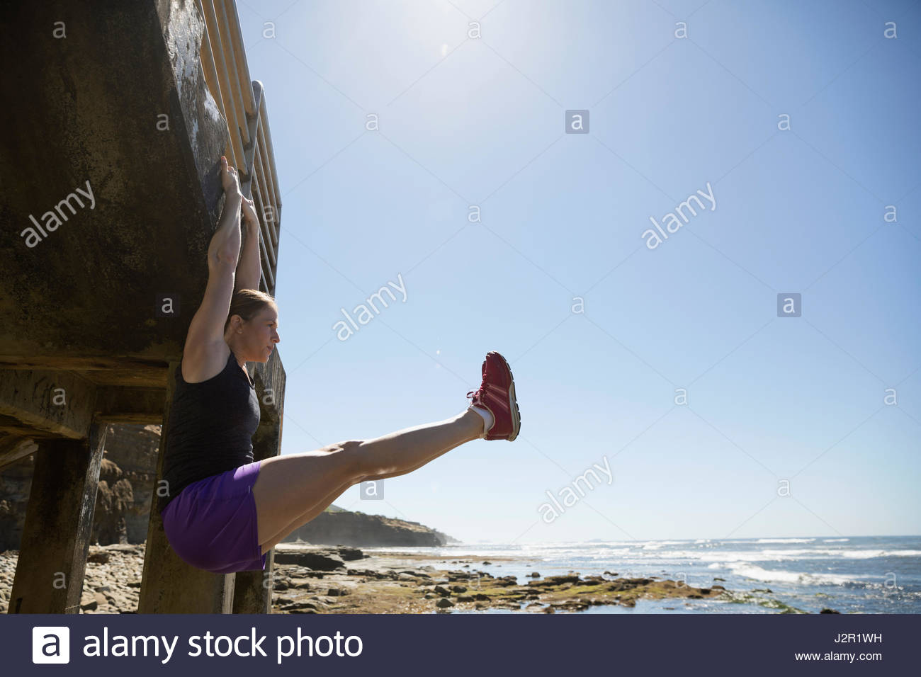 Woman exercising, hanging from stairs doing leg lifts along sunny ocean Stock Photo