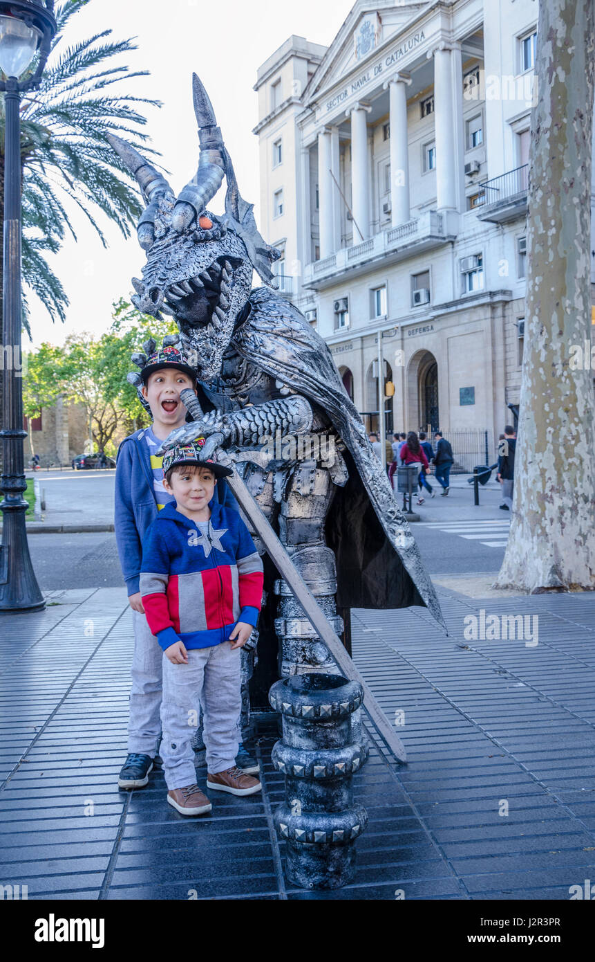 A couple of young brothers pose for a photo with a street performer dressed as a silver dragon on La Rambla in Barcelona, Spain. Stock Photo