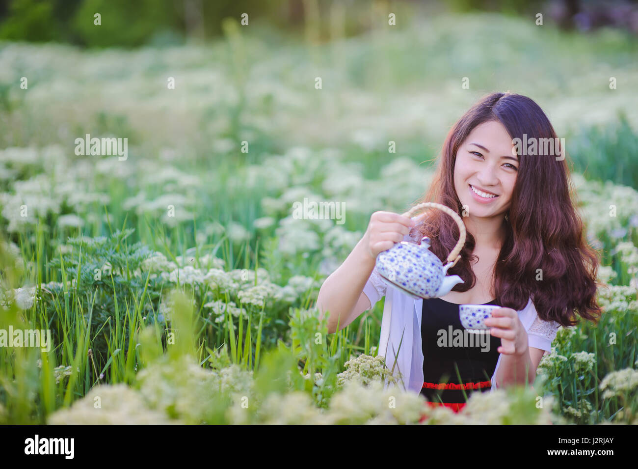 Young Chinese smiling girl pouring tea outdoors. Asian beauty posing for photos, holding small tea pot. Pretty girl sitting in flowering meadow, grass Stock Photo