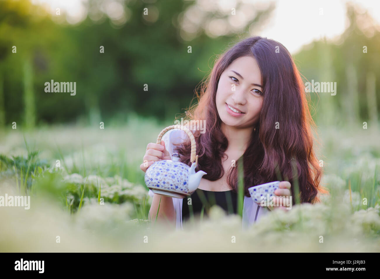 Young Chinese smiling girl pouring tea outdoors. Asian beauty posing for photos, holding small tea pot. Pretty girl sitting in flowering meadow, grass Stock Photo