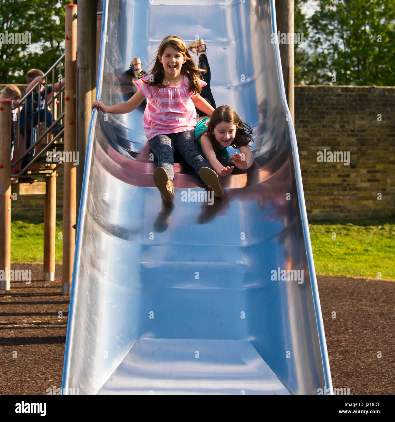 Vertical portrait of two girls sliding down a slide together in the sunshine. Stock Photo
