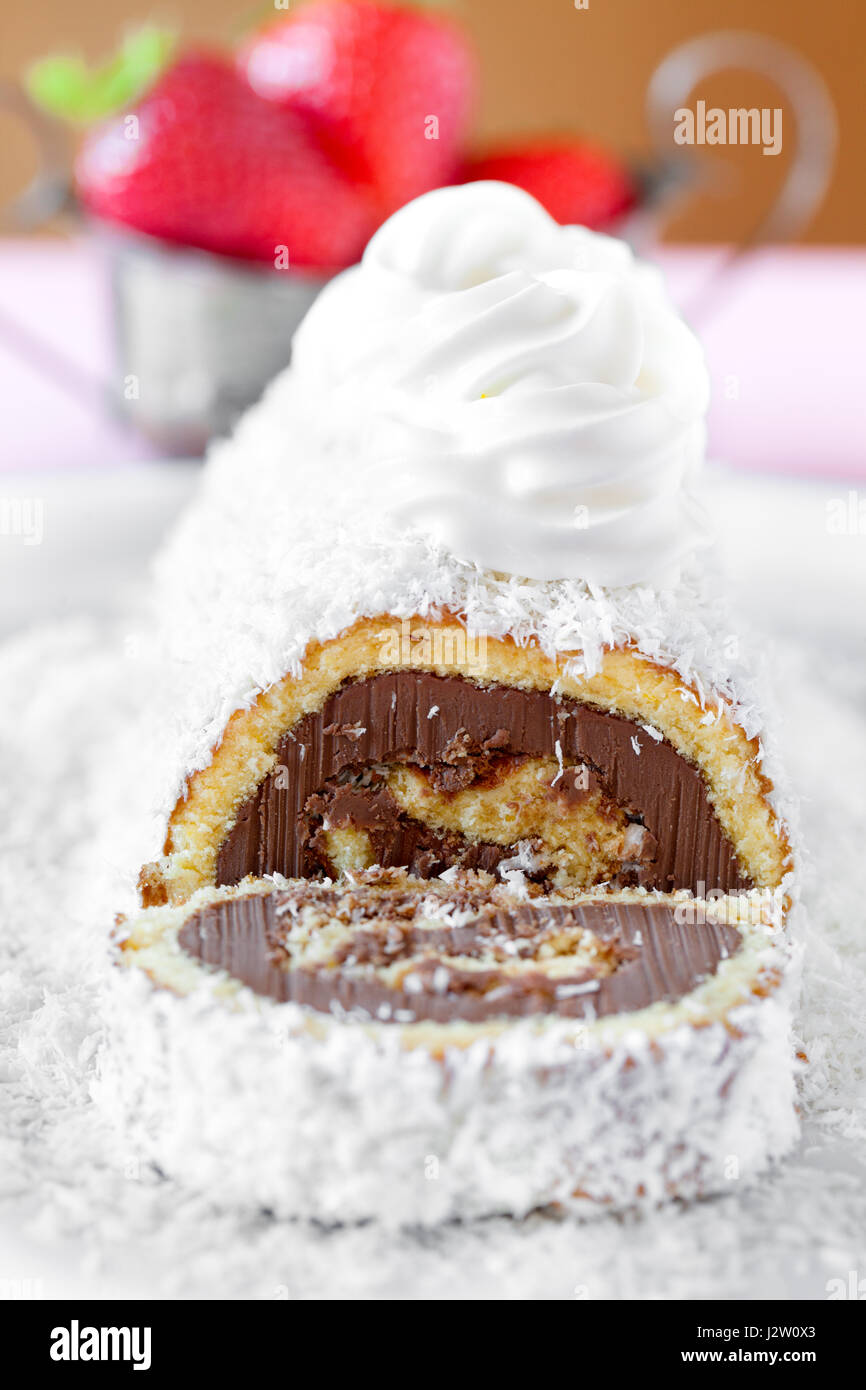 Cake roll of chocolate with coconut decorated with whipped cream, with strawberries in the background. Stock Photo