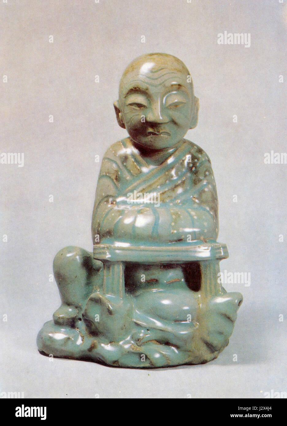 This Celadon ceramic piece features a seated Arhat, an important figure in Buddhist tradition, decorated with underglaze white slip, representing craftsmanship in Chinese pottery. Stock Photo