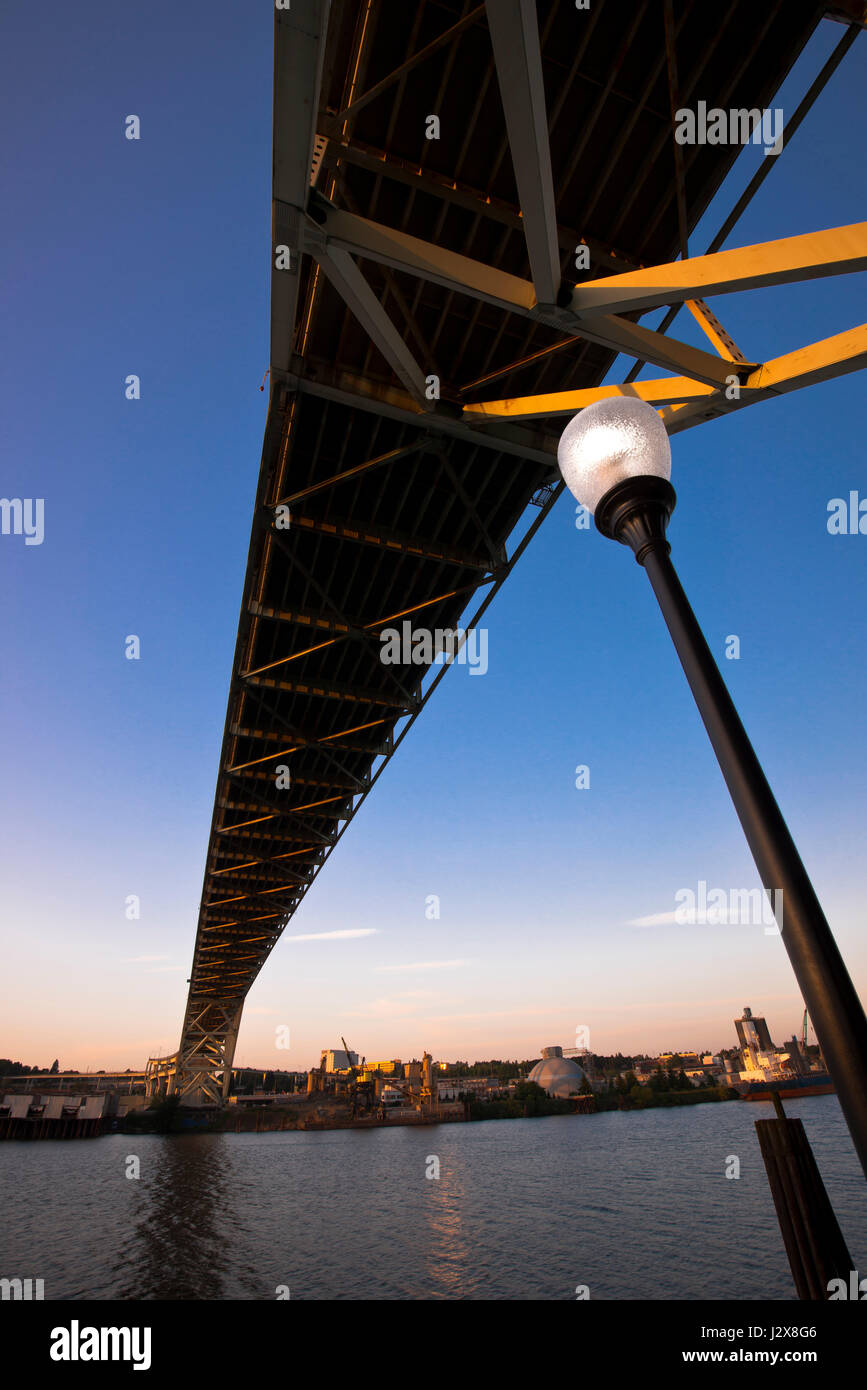 Modern perspective on the bridge over the river and city street lamp against the blue evening sky and sunlit industrial area on beach with reflection Stock Photo