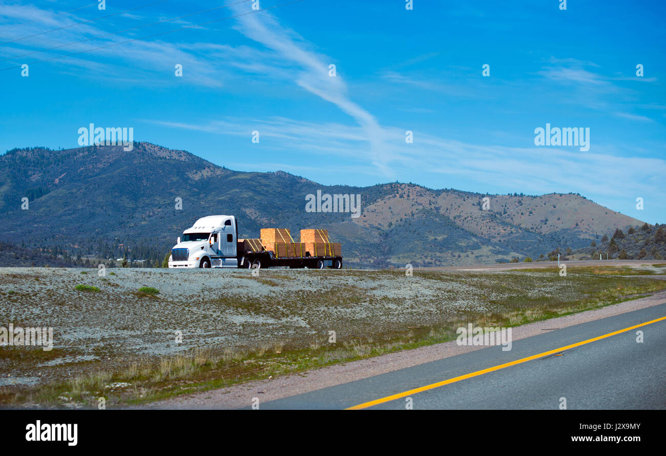 White powerful modern professional semi truck to haul a trailer carrying packaged bundles of wooden bars on interstate highway in California among bal Stock Photo