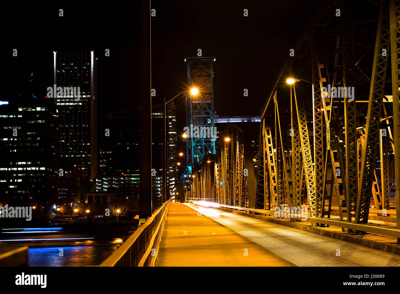 Evening lights lit metal drawbridge with two supporting towers across the river on a background of a passing downtown city blurred silhouette Stock Photo