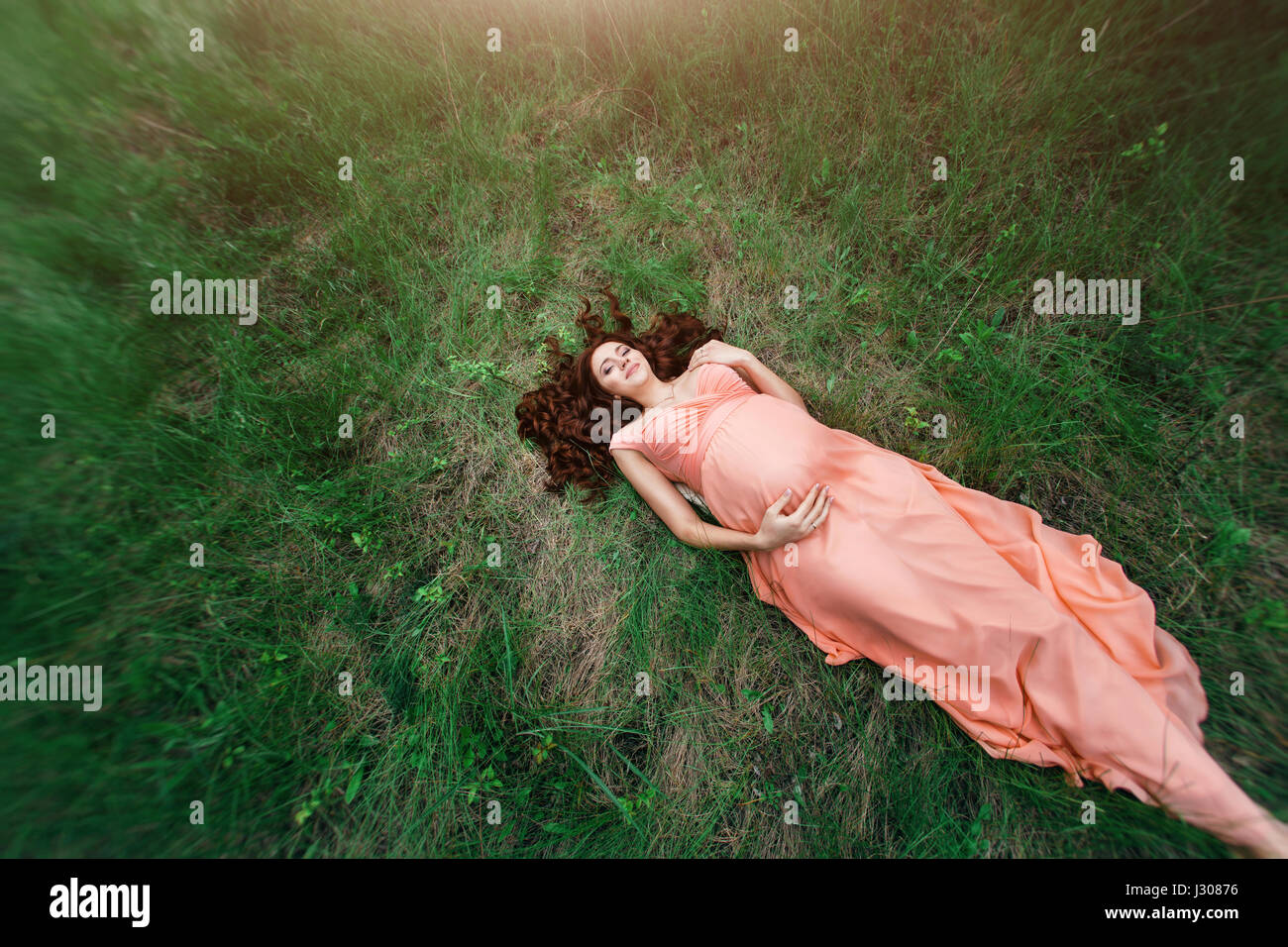 Goorgeous woman laying on green grass field in summer. Attractive mcaucasian girl in salmon dress resting on nature. Top view creative shot on fisheye Stock Photo