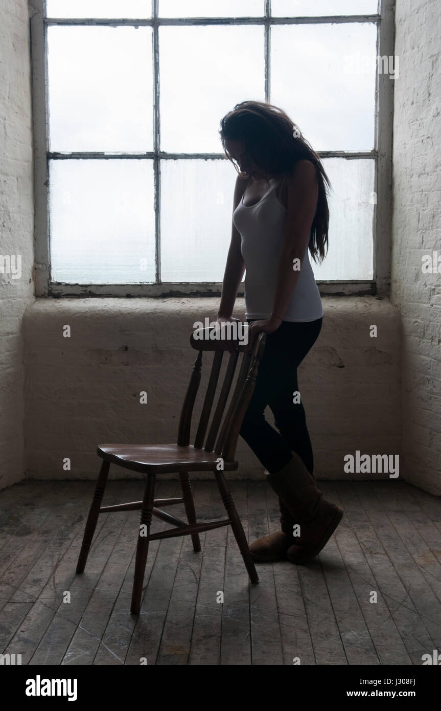 Sad young woman leaning on a chair standing near the window Stock Photo