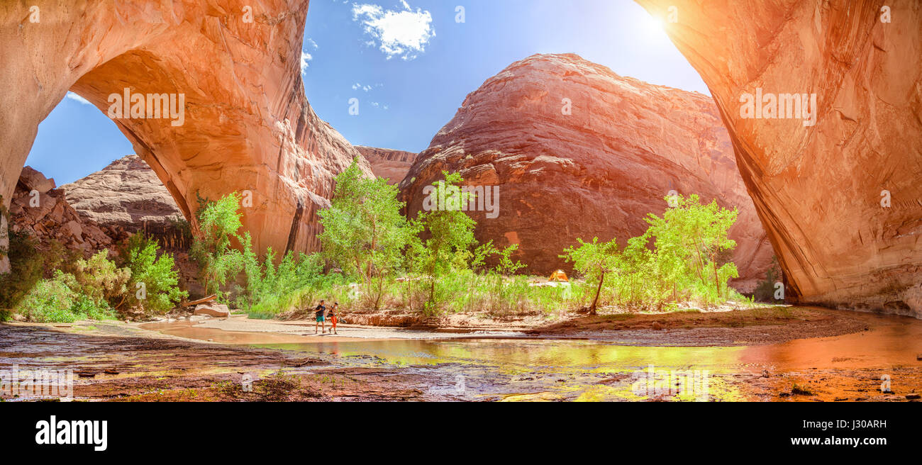 Wide angle view of two hikers backpacking beneath stunning Jacob Hamblin Arch in Coyote Gulch on a sunny day with blue sky and clouds in summer, Grand Stock Photo