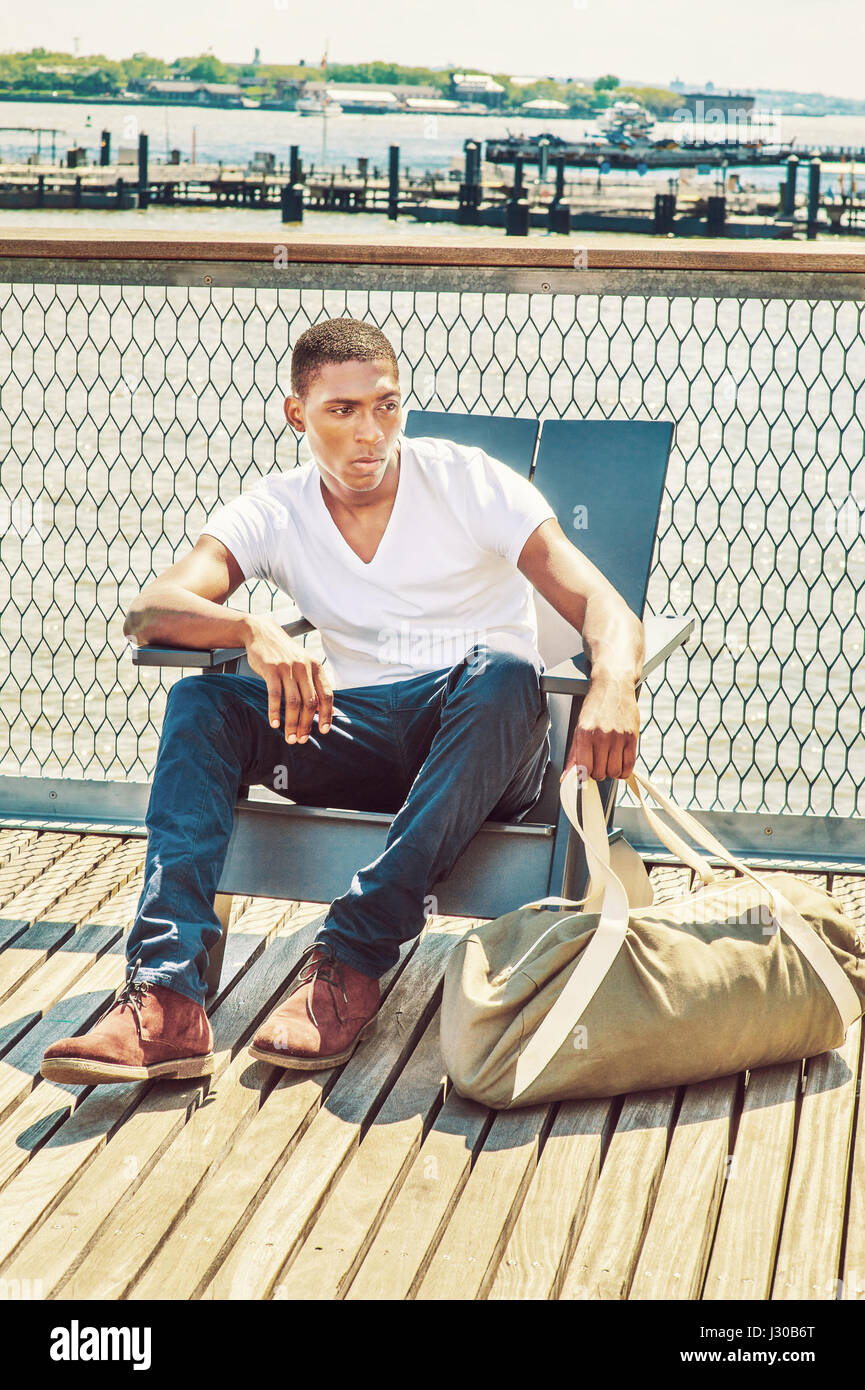 Traveling. Wearing white, V neck T shirt, blue pants, brown boot shoes, a  hand carrying duffel bag, a young black college student is sitting on dock  b Stock Photo - Alamy