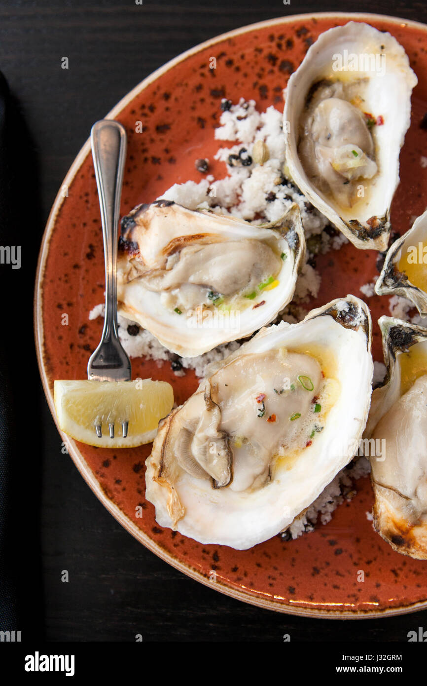 Overhead shot of grilled oysters with butter and lemon, on an orange plate and dark surface Stock Photo