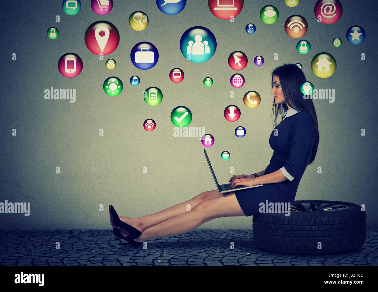 Young woman sitting on a tire working on laptop computer with social media application icons flying out of screen. Modern technology and people concep Stock Photo