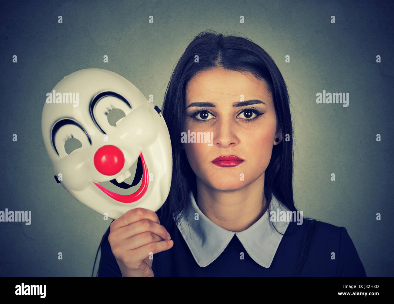 Young woman with sad face expression holding clown mask expressing cheerfulness happiness isolated on gray wall background Stock Photo