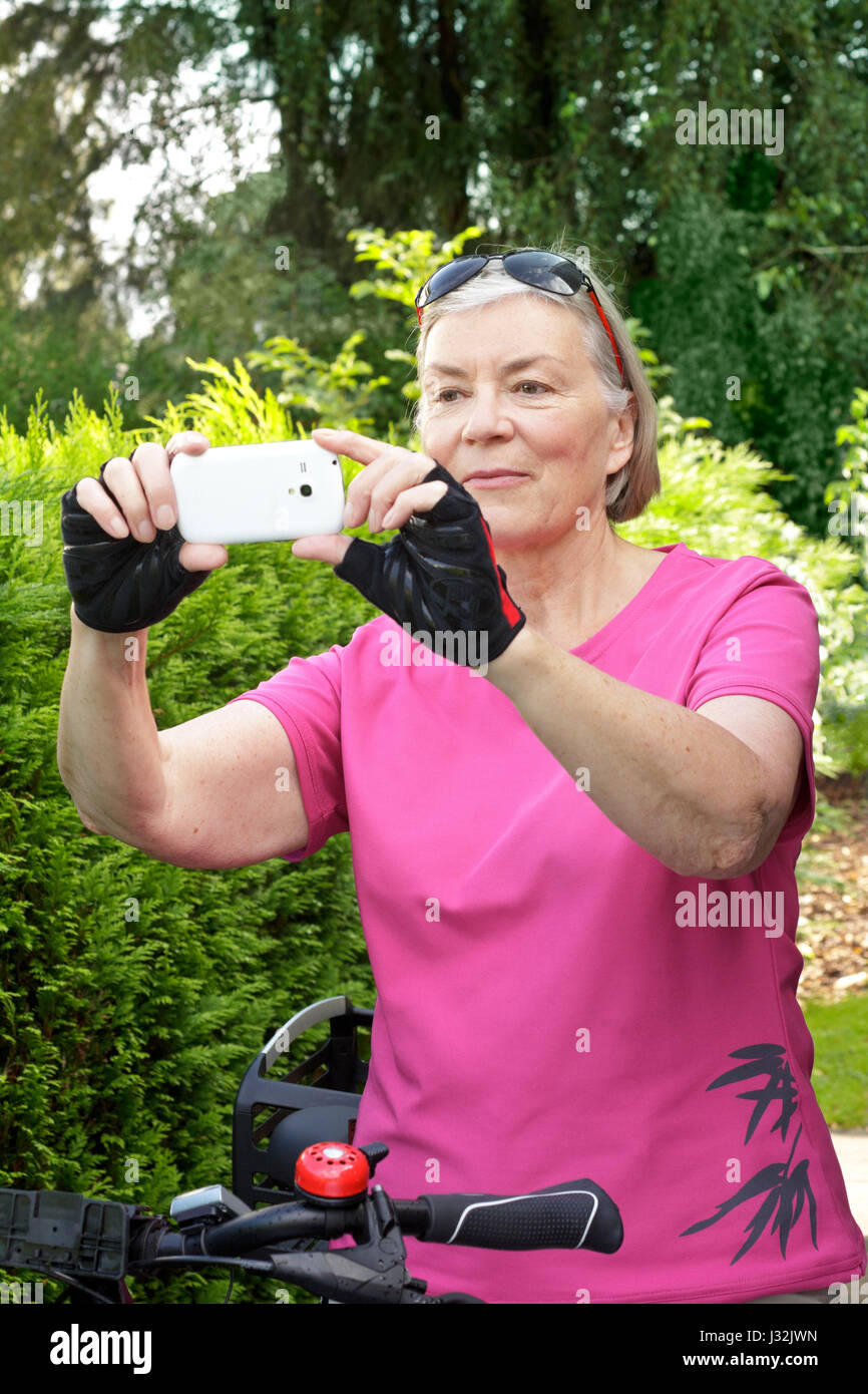 Senior woman on a bicycle tour in summer wearing protective goves, using her cell phone to take pictures of the landscape, green background Stock Photo
