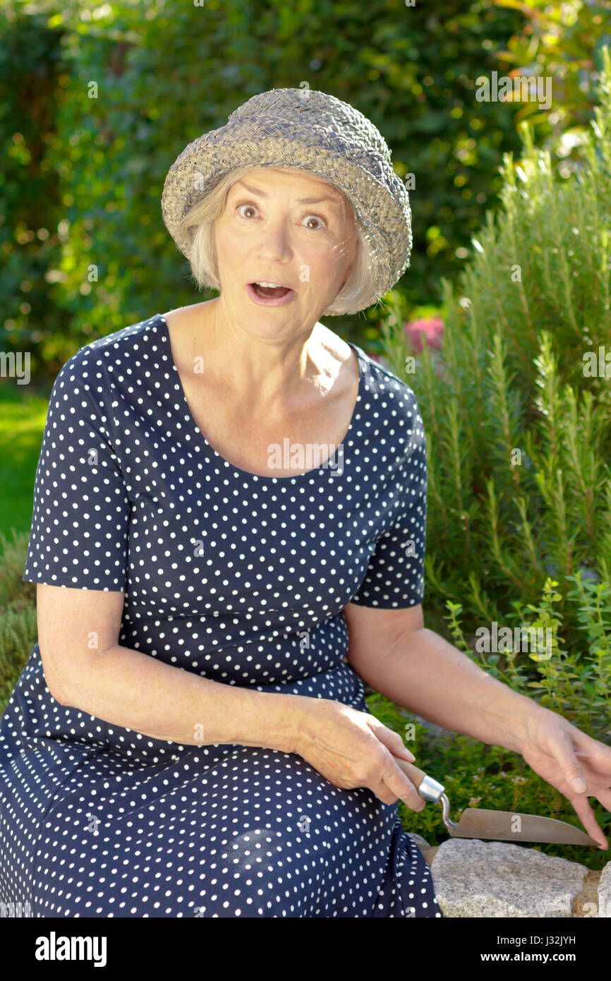Senior white woman in a blue polka dotted and straw hat in her garden yard showing an exaggerated surprise, joy in garden concept Stock Photo