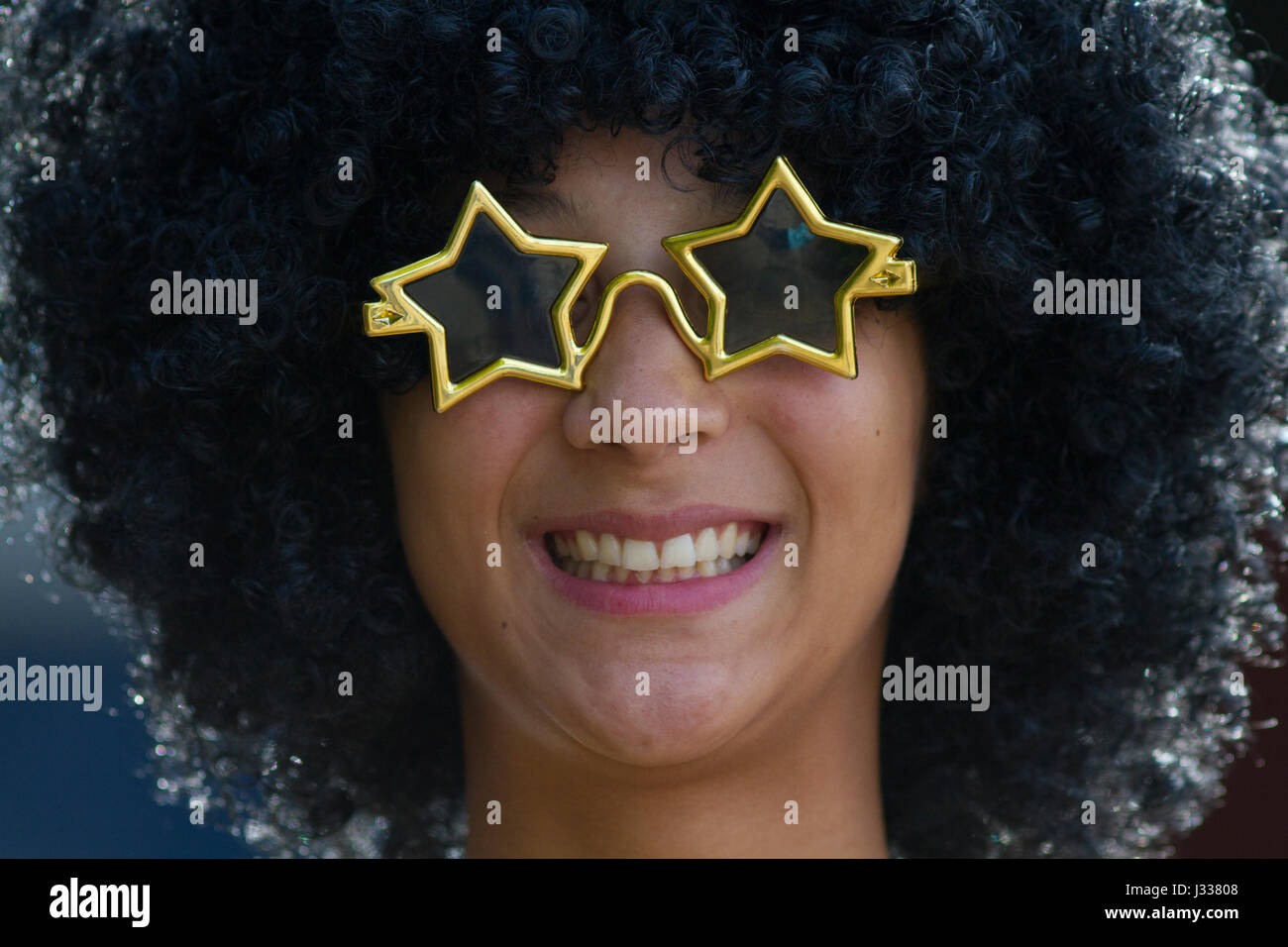 Mixed race teenage girl smiling in Sixties fancy dress with star shaped sunglasses and an afro wig, Sussex, Uk Stock Photo