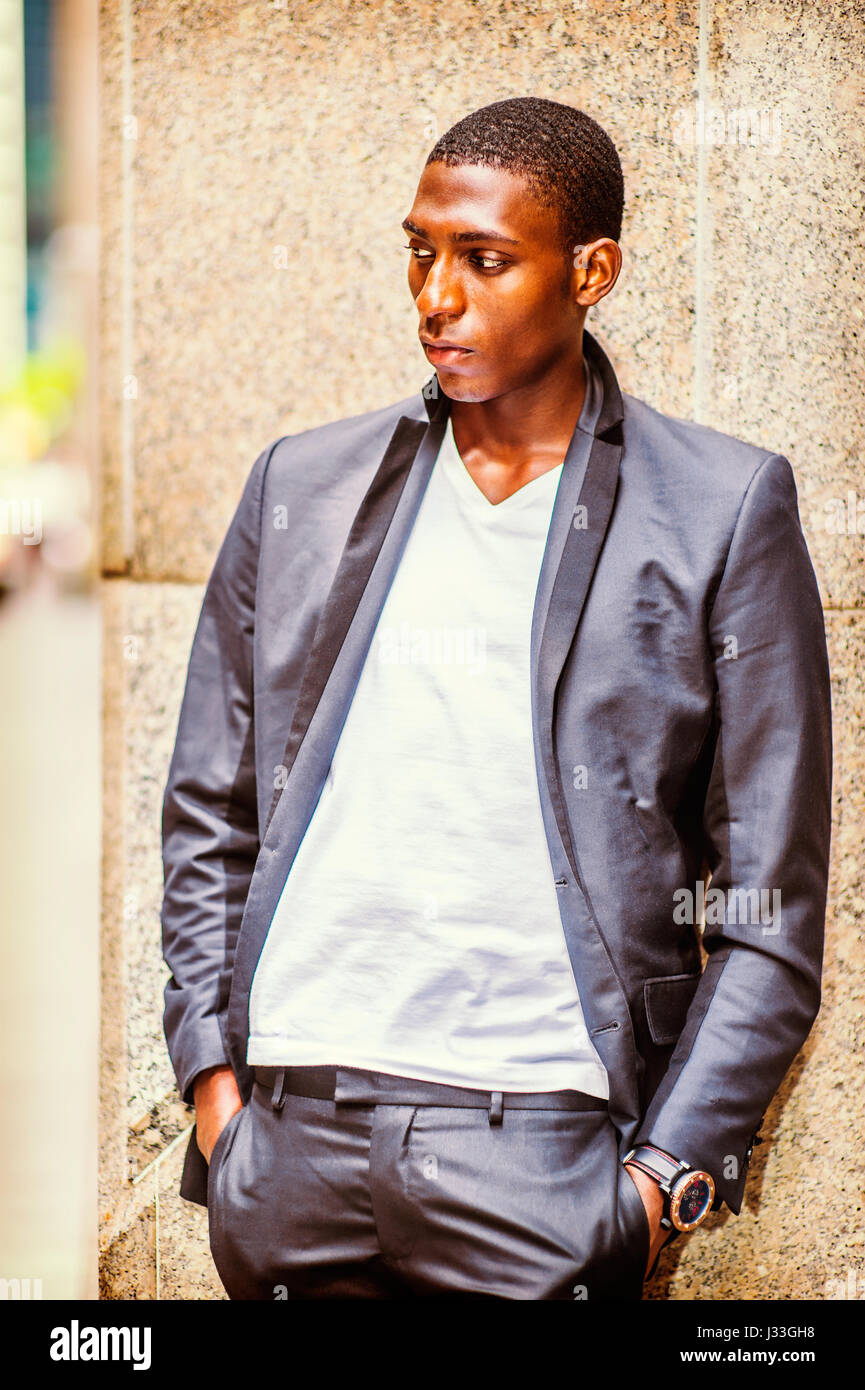 Portrait of African American Teenage Boy in New York, wearing black fashionable jacket, white v neck shirt, standing against column on street, thinkin Stock Photo