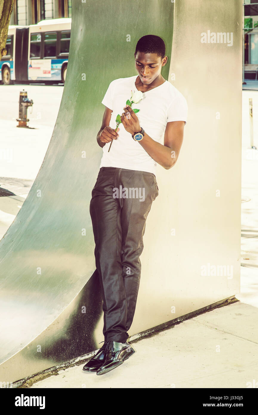 Young African American Man missing you, wearing white V neck T shirt, black pants, leather shoes, standing against metal structure on street in New Yo Stock Photo