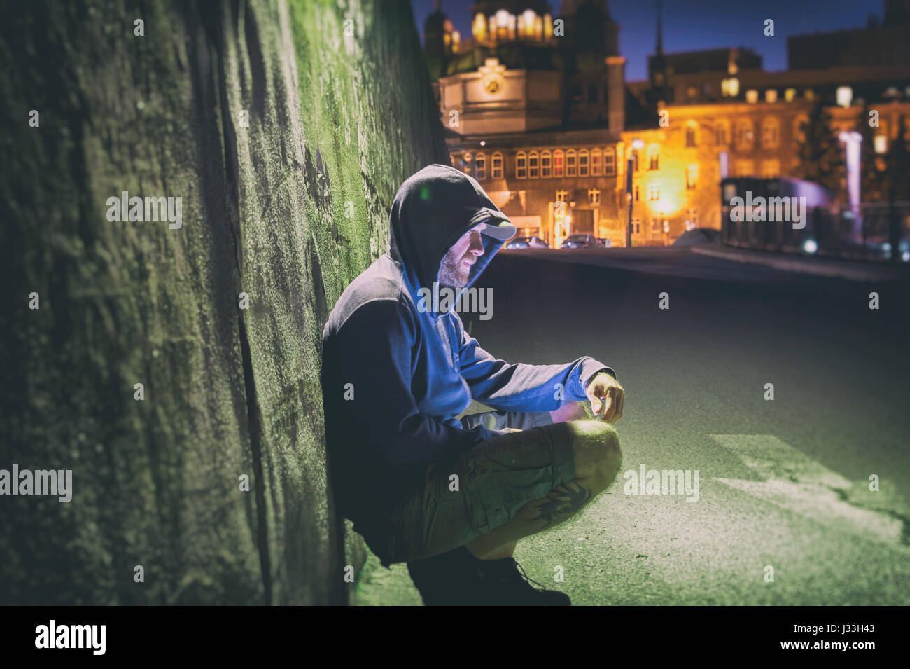 Sad and lonely man with hoodie sitting against a wall at night Stock Photo