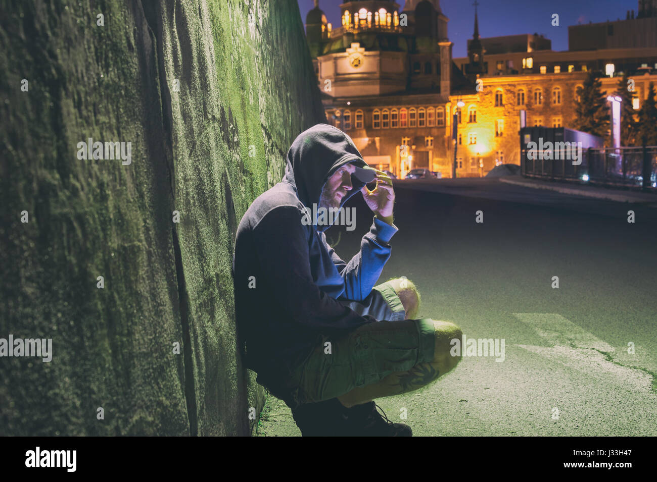 Sad and lonely man with hoodie sitting against a wall at night Stock Photo
