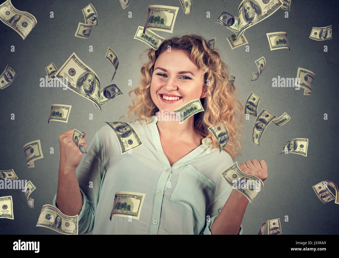 Portrait happy woman exults pumping fists celebrates success under money rain falling down dollar bills banknotes isolated on gray wall background Stock Photo