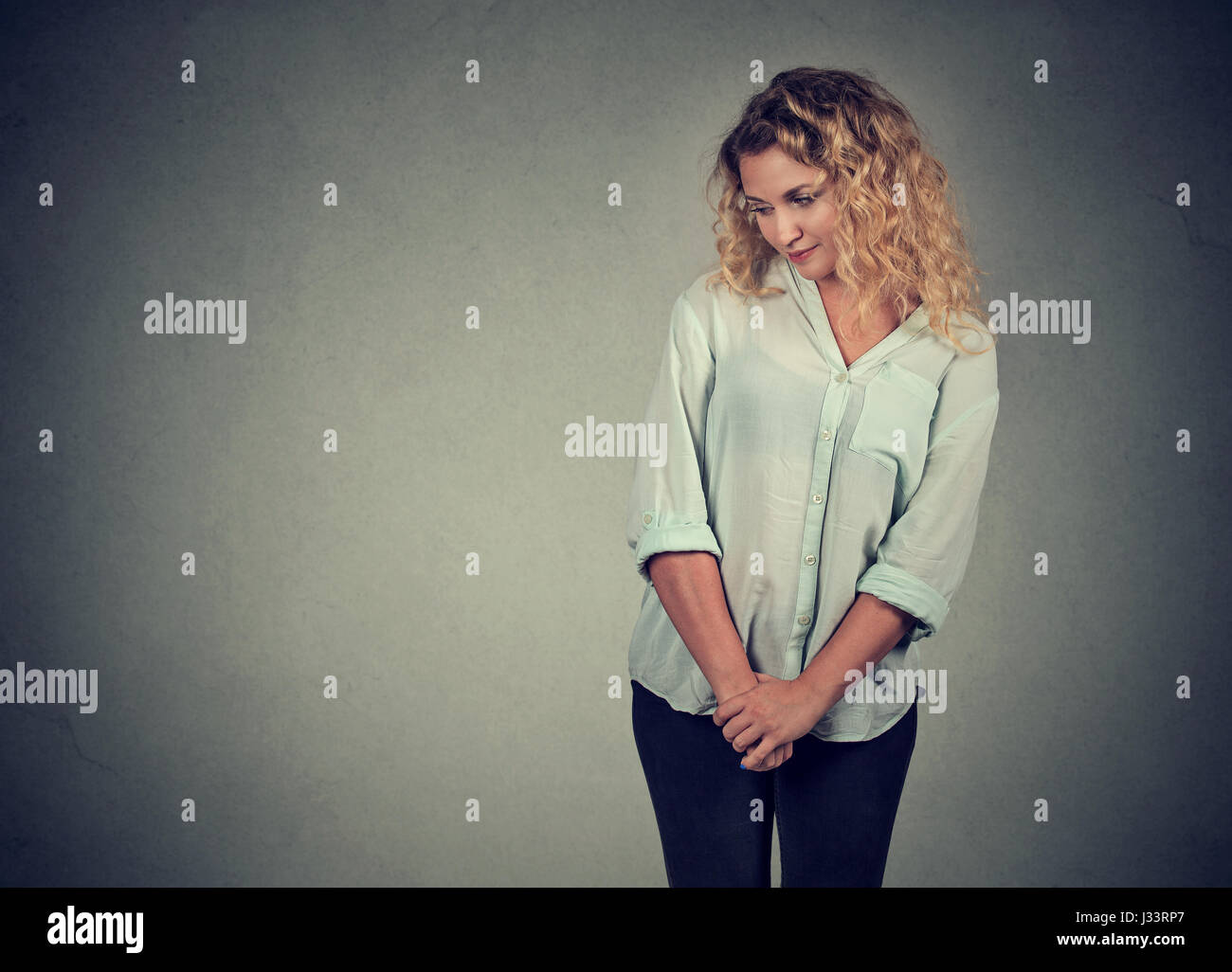 Shy insecure young woman looking down avoiding eye contact standing isolated on gray wall background Stock Photo