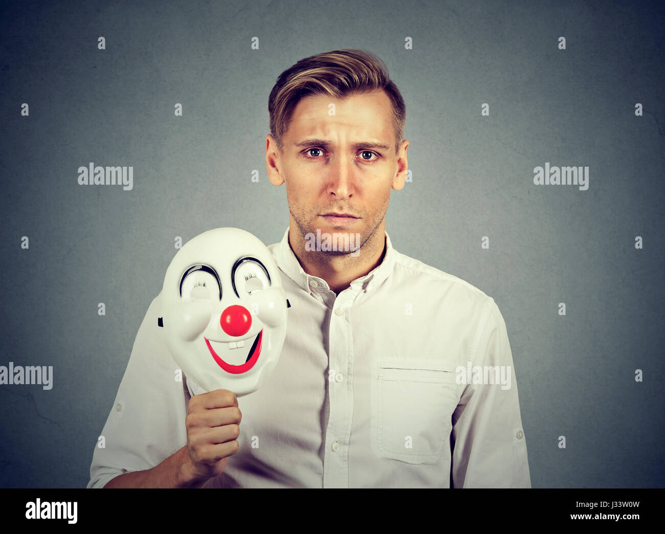 Young sad man with happy clown mask isolated on gray wall background. Human emotions Stock Photo