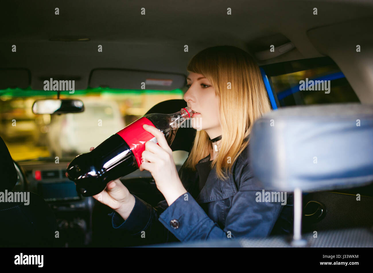 portrait of blonde woman drinking from large drink bottle, quenching thirst, sitting in car in back seat, photo is taken from rear of vehicle Stock Photo