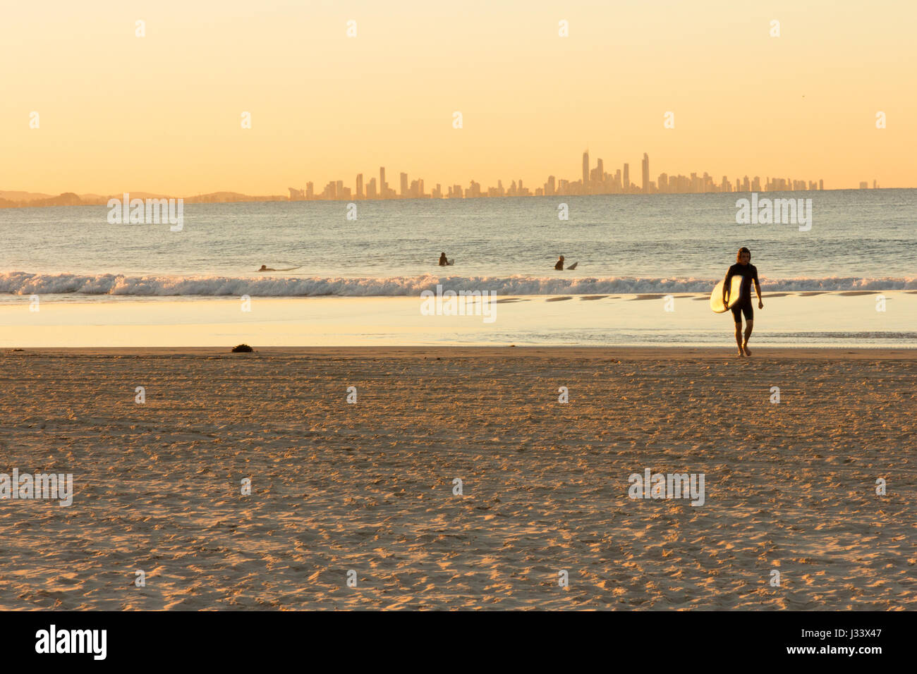 Surfer walks away from the sea on Coolangatta beach at sunset with Surfer's Paradise in the background Stock Photo