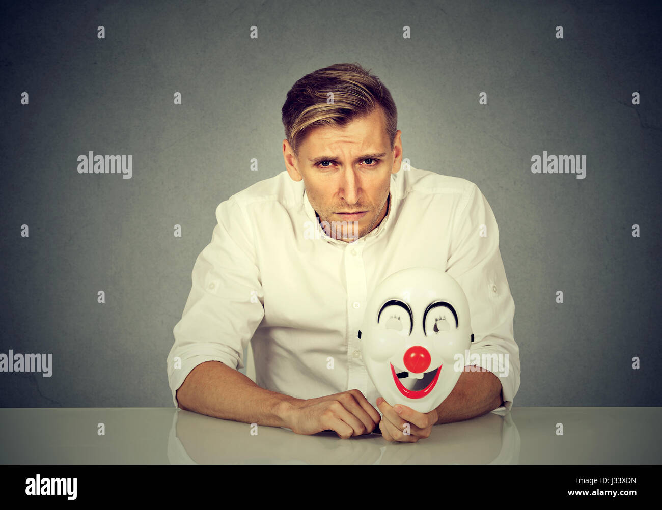 Portrait young upset worried man with sad expression holding clown mask expressing cheerfulness happiness isolated on gray wall background. Human emot Stock Photo