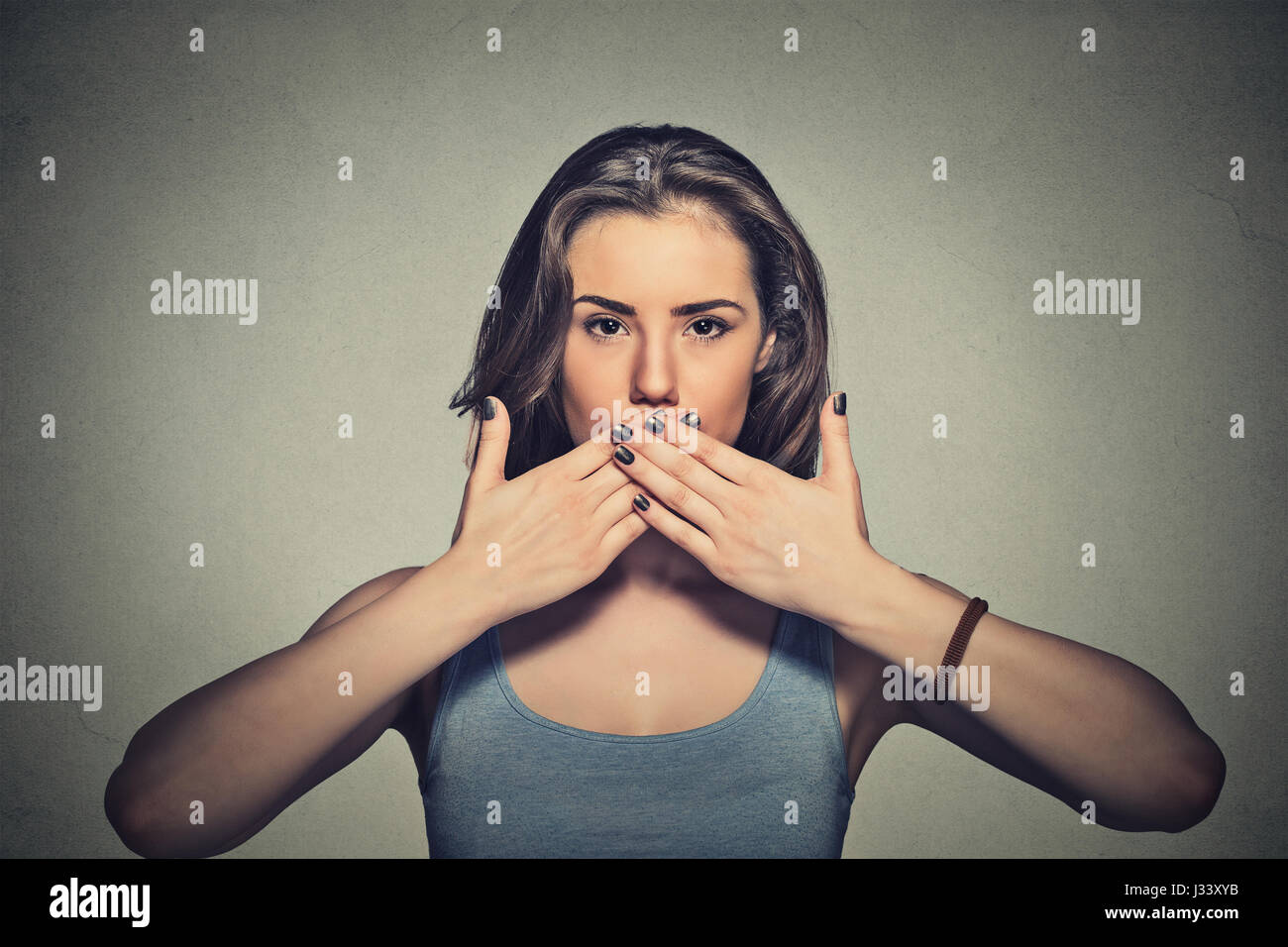 Closeup portrait of young woman covering with hands her mouth isolated on gray wall background Stock Photo