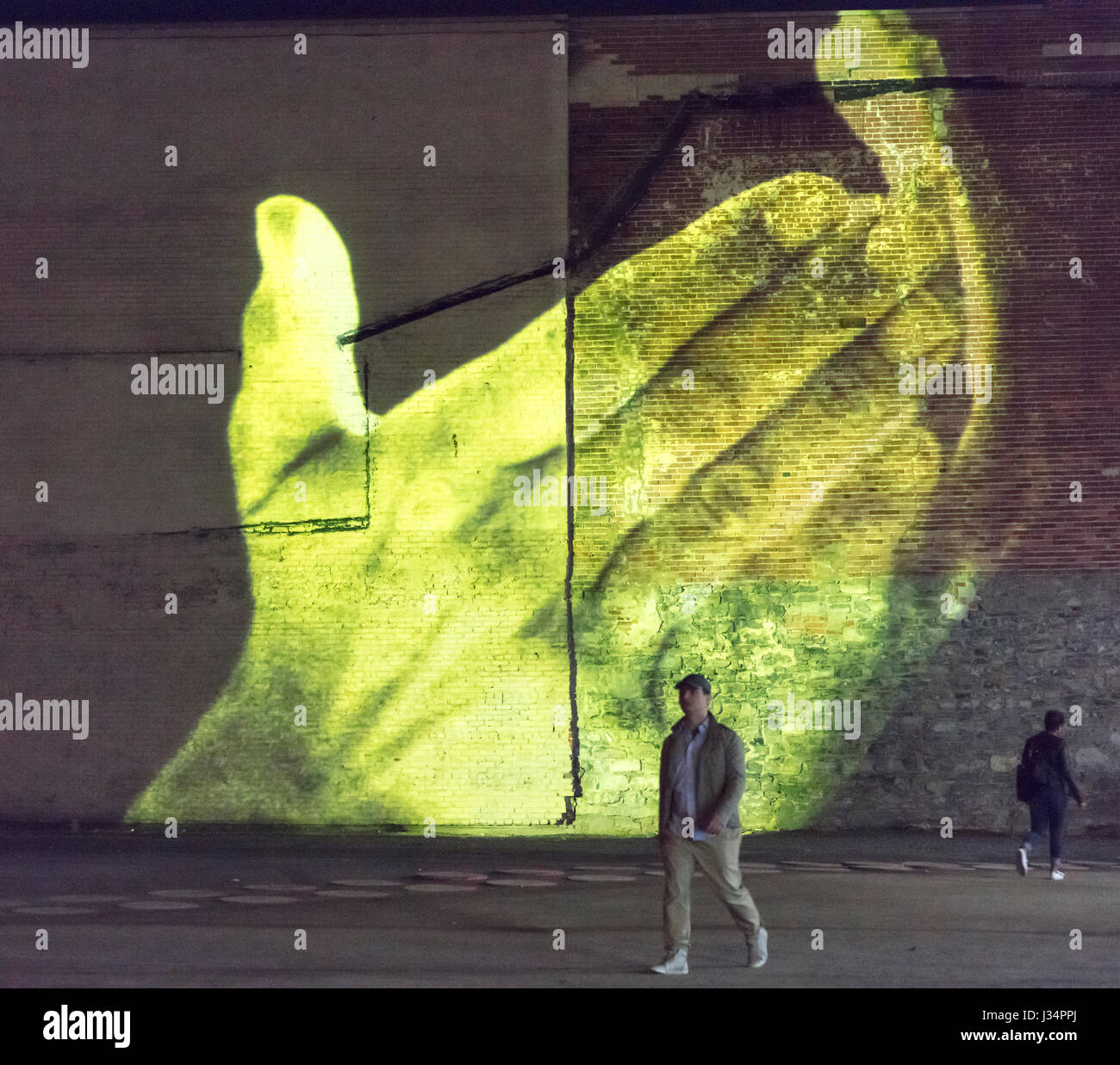 Boulevard St Laurent, Montreal, Quebec, Canada - 27 April 2017: pedestrians walk in front of video of yellow hands projected on brick wall Stock Photo
