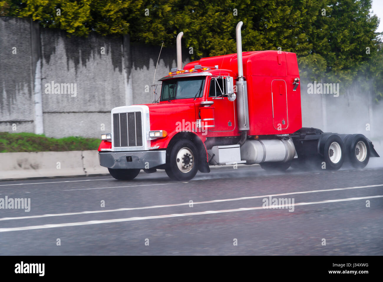 Professional classical bonnet red semi-truck with a long cab and vertical exhaust pipes drive on highway with multiple lanes, raising by wheels Stock Photo