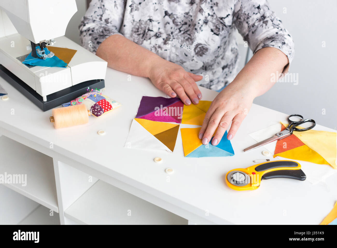 needlework and quilting in the workshop of a woman tailor - hands of tailor women put on the desktop the pieces of colored fabric, lay next to the scissors, buttons, pins and threads. Stock Photo