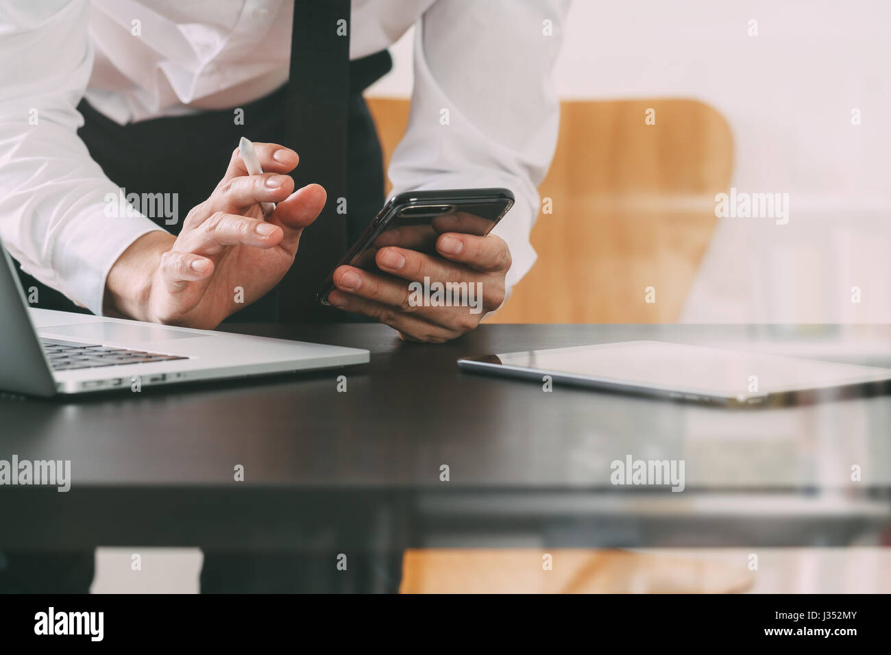 close up of businessman working with smart phone and digital tablet and laptop computer on wooden desk in modern office with glass reflected view Stock Photo