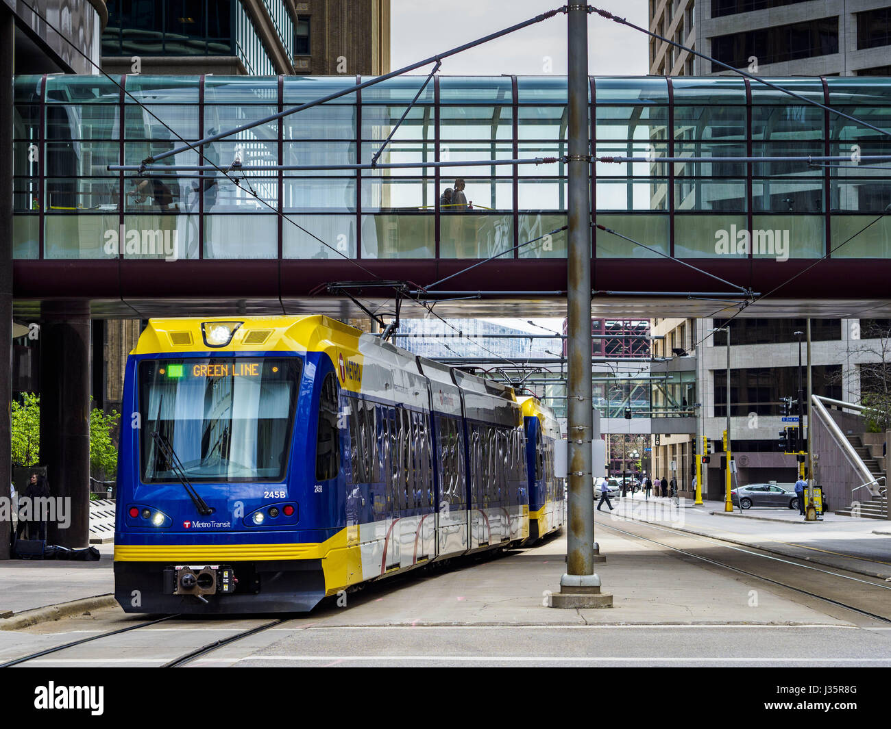 Minneapolis, Minnesota, USA. 3rd May, 2017. A Minneapolis light rail train goes under the skyway on 5th Street in Minneapolis. The skyways are enclosed pedestrian overpasses that connect downtown buildings. The Minneapolis Skyway was started in the early 1960s as a response to covered shopping malls in the suburbs that were drawing shoppers out of the downtown area. The system grew sporadically until 1974, when the construction of the IDS Center and its center atrium, called the Crystal Court, served as a hub for the downtown skyway system. There are 8 miles of skyways, connecting most of the Stock Photo