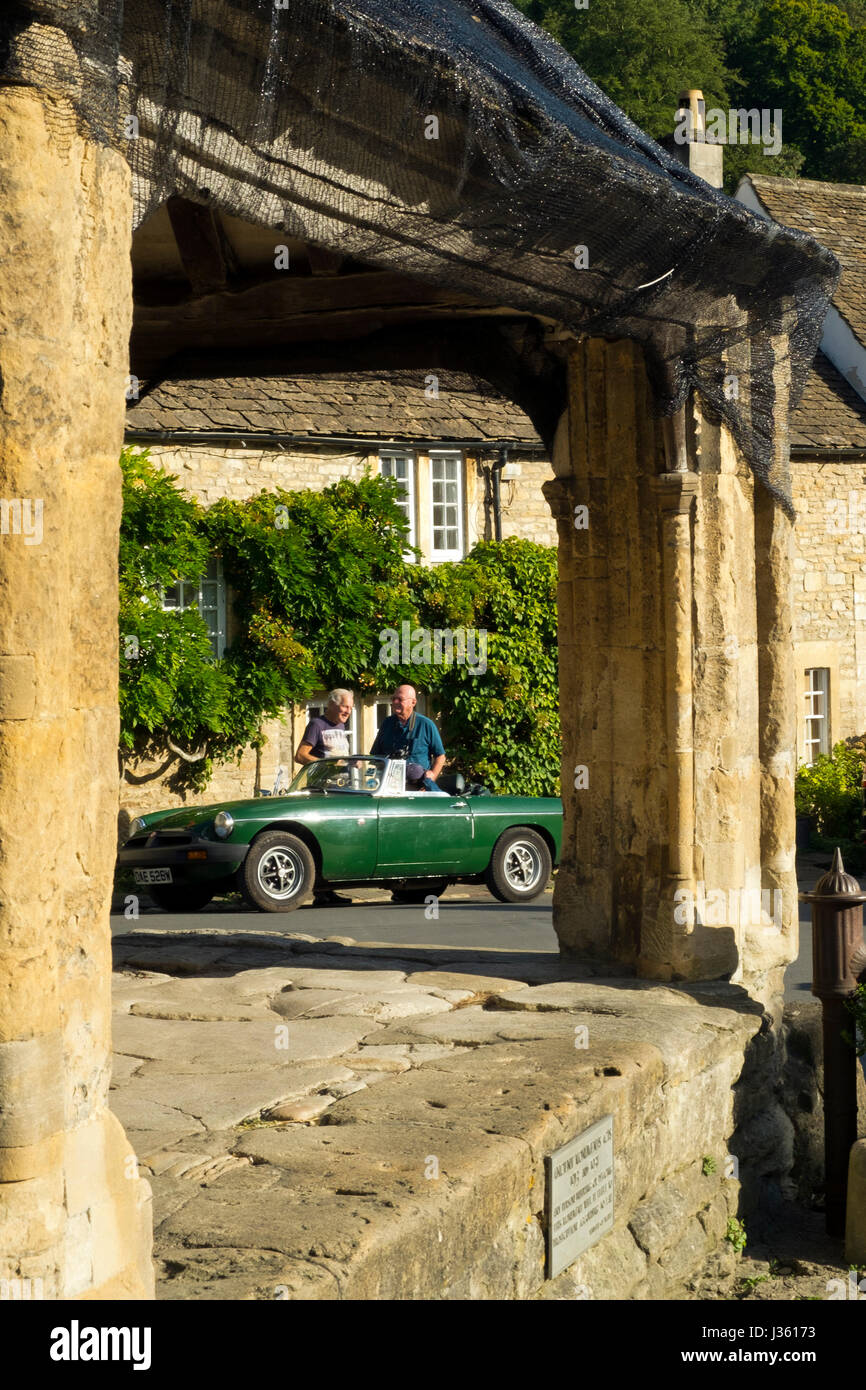 Two gentlemen stand discussing a classic MGB sports car near the 14thC Market Cross in Castle Combe, The Cotswolds, Wiltshire, UK Stock Photo