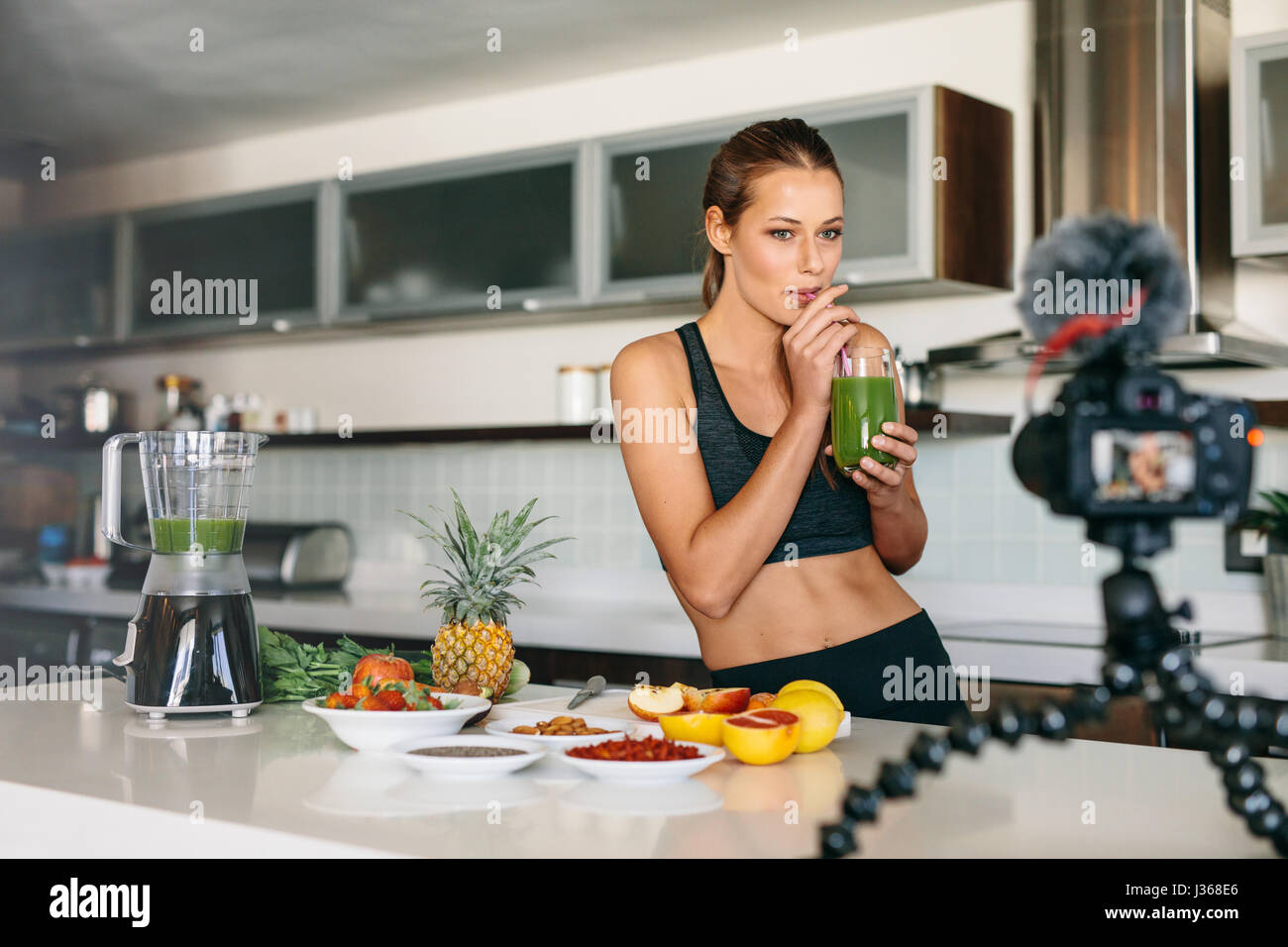 Young lady drinking green juice at the kitchen table looking at a camera mounted on tripod. Woman preparing a healthy breakfast with fruits and vegeta Stock Photo