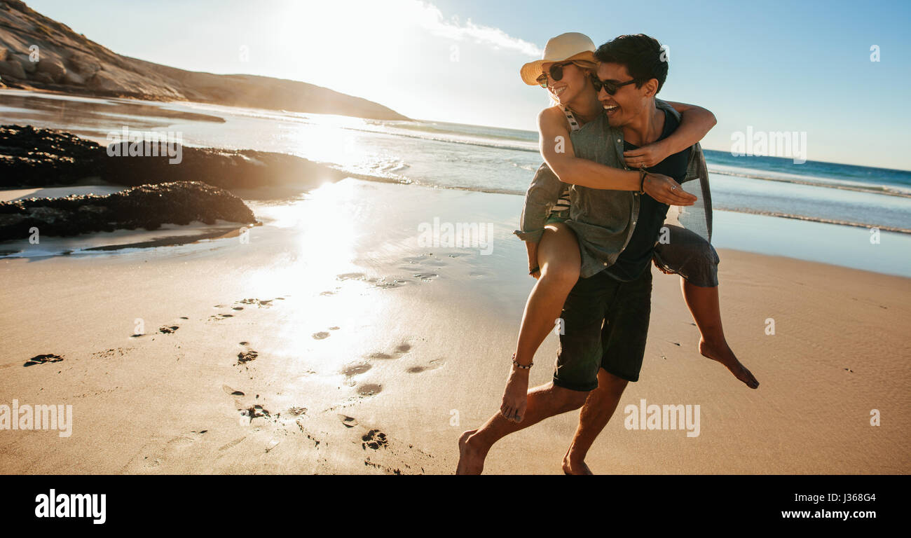 Man giving piggyback ride to girlfriend on beach. Happy young couple having fun on the seashore, enjoying summer holidays. Stock Photo