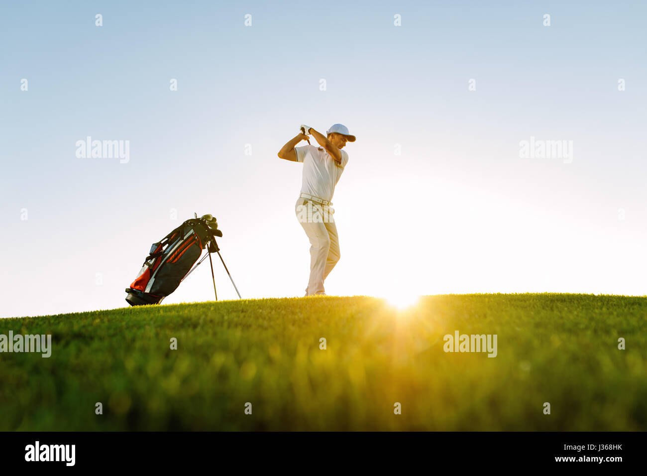 Low angle shot of male golfer taking shot while standing on field. Full length of golf player swinging golf club on sunny day. Stock Photo
