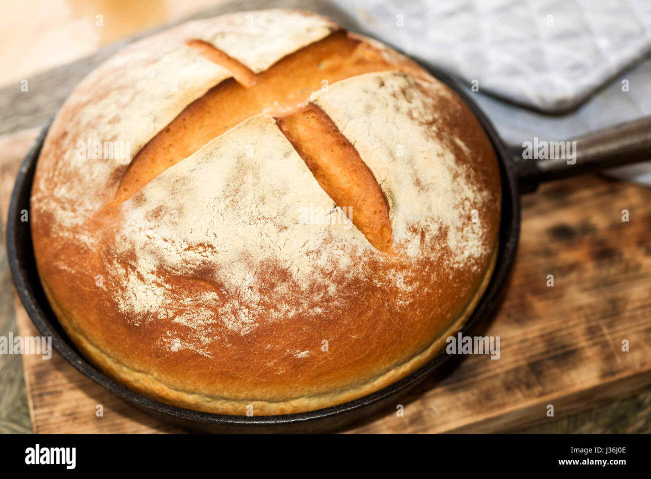 Newly baked artisan bread in cast iron skillet on wooden cutting board. Gray potholder at side. Stock Photo