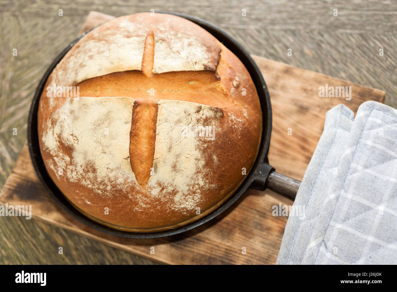 Newly baked artisan bread in cast iron skillet on wooden cutting board. Gray potholder at side. Stock Photo