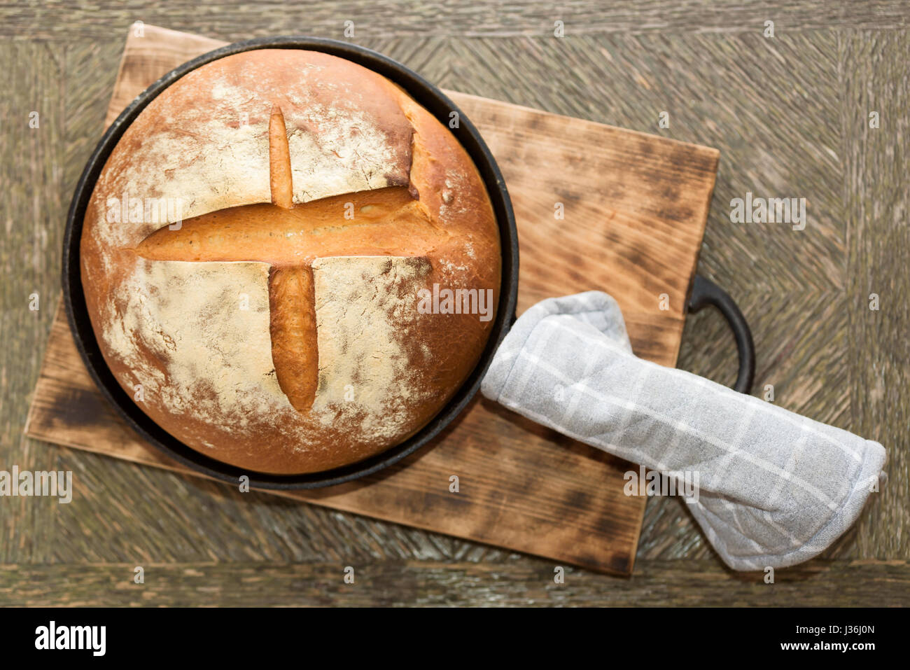 Newly baked artisan bread in cast iron skillet on wooden cutting board. Gray potholder at side. Stock Photo