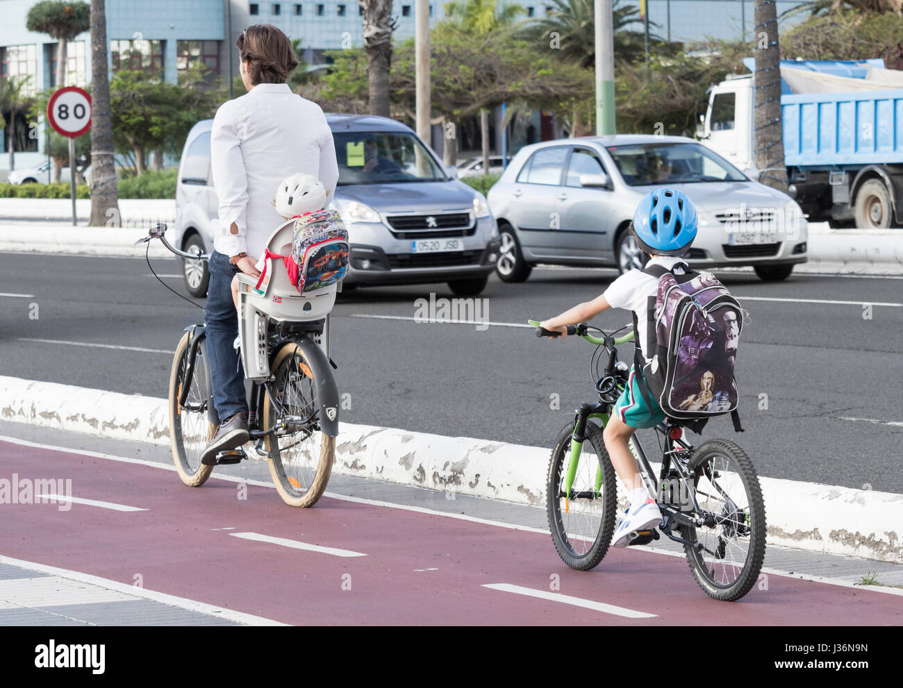 Young boy in school uniform riding to school on bicycle behind adult who is carrying small child. Stock Photo